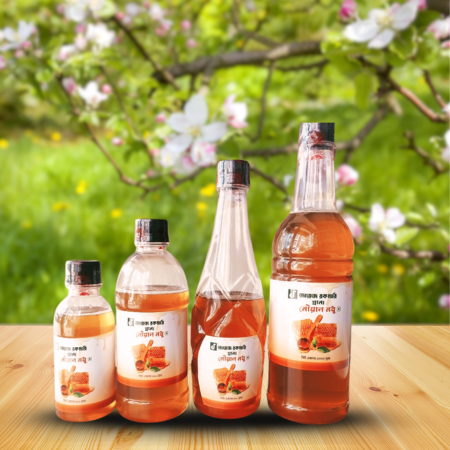 A display of four plastic bottles of natural honey labeled ""জায়ে রকমারি মধু" in varying sizes, placed on a wooden surface with a vibrant green garden background. Blooming white and pink apple blossoms on a tree branch are visible above, emphasizing the natural and organic setting.